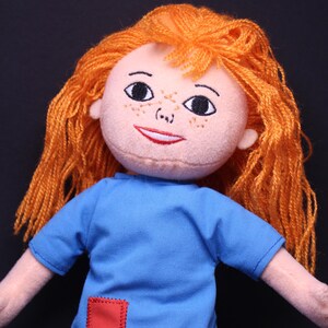 Pode incluir: Boneca feita &agrave; m&atilde;o com rosto p&ecirc;ssego claro, sardas e cabelo de fio laranja. A boneca usa uma camiseta azul com um patch quadrado vermelho. Os olhos da boneca s&atilde;o pretos e a boca vermelha.