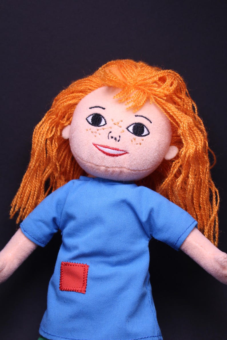 Pode incluir: Boneca feita &agrave; m&atilde;o com rosto p&ecirc;ssego claro, sardas e cabelo de fio laranja. A boneca usa uma camiseta azul com um patch quadrado vermelho. Os olhos da boneca s&atilde;o pretos e a boca vermelha.