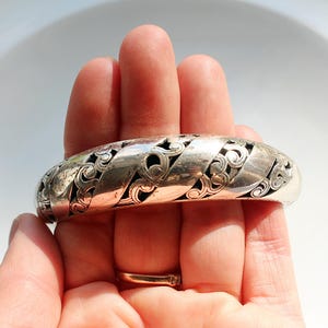 Puede incluir: Un brazalete plateado con un dise&ntilde;o calado intrincado. El brazalete presenta una serie de bandas entrelazadas con patrones florales en espiral. El brazalete se sostiene en una mano, mostrando su superficie pulida y su detallada artesan&iacute;a.