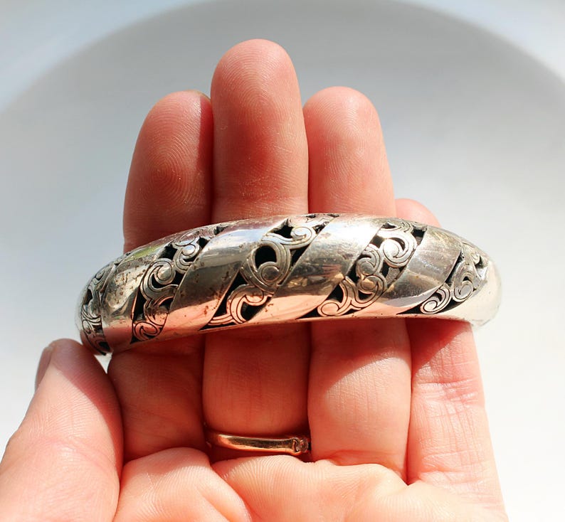Puede incluir: Un brazalete plateado con un dise&ntilde;o calado intrincado. El brazalete presenta una serie de bandas entrelazadas con patrones florales en espiral. El brazalete se sostiene en una mano, mostrando su superficie pulida y su detallada artesan&iacute;a.