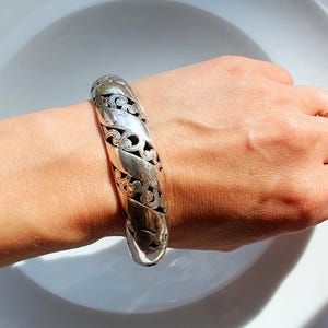 Puede incluir: Un brazalete plateado con un intrincado dise&ntilde;o calado. El brazalete se lleva en la mu&ntilde;eca y presenta un patr&oacute;n repetido de formas arremolinadas y detalles recortados. El brazalete mide aproximadamente 5 cm de ancho.