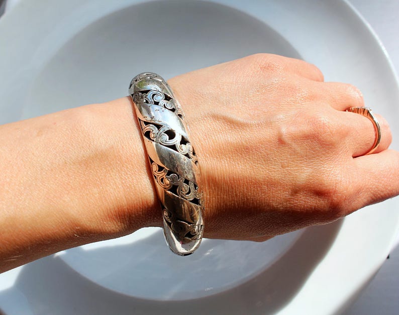 Puede incluir: Un brazalete plateado con un intrincado dise&ntilde;o calado. El brazalete se lleva en la mu&ntilde;eca y presenta un patr&oacute;n repetido de formas arremolinadas y detalles recortados. El brazalete mide aproximadamente 5 cm de ancho.