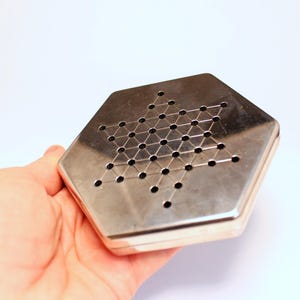 Puede incluir: Un tablero de juego de solitario plateado con un patrón de agujeros en forma de estrella. El tablero hexagonal tiene un acabado metálico brillante. El tablero de juego se sostiene en una mano sobre un fondo blanco.