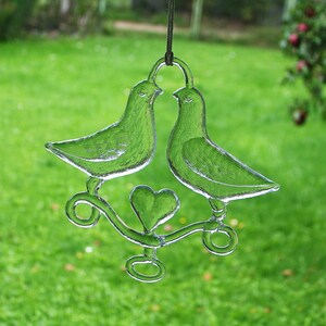 Puede incluir: Un adorno de vidrio con dos palomas posadas en una rama con un coraz&oacute;n en el centro. Las palomas son de vidrio transparente y la rama es de vidrio esmerilado.