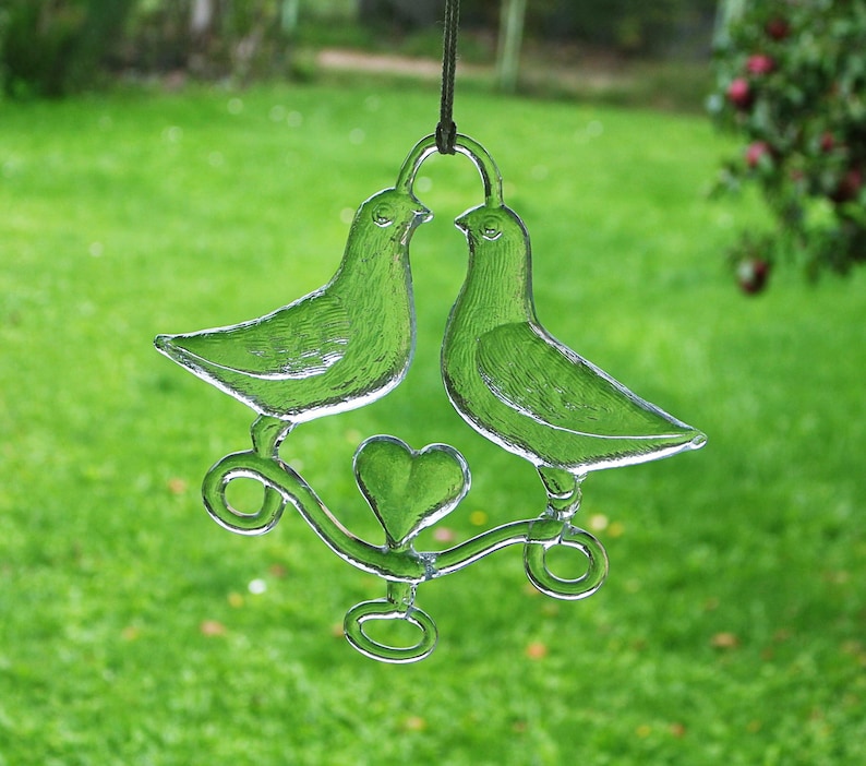Puede incluir: Un adorno de vidrio con dos palomas posadas en una rama con un coraz&oacute;n en el centro. Las palomas son de vidrio transparente y la rama es de vidrio esmerilado.