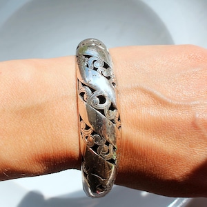 Puede incluir: Brazalete plateado con dise&ntilde;o calado. El brazalete presenta un patr&oacute;n repetitivo de formas curvas y espirales. El metal tiene un aspecto ligeramente envejecido. El brazalete se lleva en la mu&ntilde;eca.