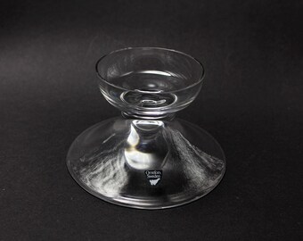 May include: A clear glass candle holder with a round base and a smaller bowl-shaped top. The candle holder has a stem connecting the base and the top. The base has a diameter of approximately 13 cm.
