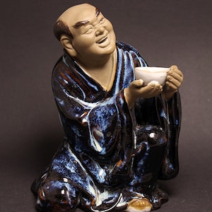Puede incluir: Figura de cerámica de un hombre calvo sonriente con una túnica azul y marrón, sosteniendo una pequeña taza blanca. La figura está arrodillada, con una expresión serena. La túnica tiene un acabado brillante y moteado.