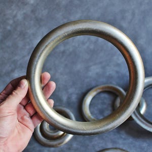 Puede incluir: Varios anillos met&aacute;licos grandes. Uno es sostenido por una mano, con otros de diferentes tama&ntilde;os en el fondo. Los anillos tienen una forma lisa y redondeada, con una apariencia ligeramente envejecida.