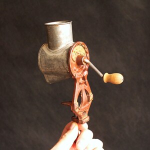 May include: An antique, hand-cranked food grinder. The grinder features a silver-colored metal hopper and grinding mechanism, with a red and gold-colored cast iron frame and a wooden handle. The grinder is designed to clamp onto a table or counter.