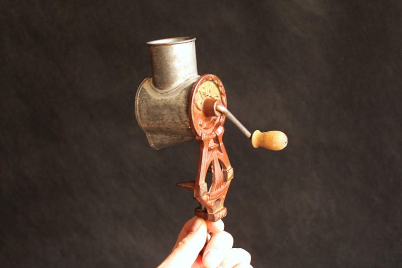 May include: An antique, hand-cranked food grinder. The grinder features a silver-colored metal hopper and grinding mechanism, with a red and gold-colored cast iron frame and a wooden handle. The grinder is designed to clamp onto a table or counter.