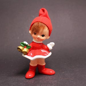 Peut inclure: Figurine de Noël en céramique représentant un enfant vêtu d'une tenue rouge avec une bordure blanche. La figurine porte un chapeau rouge, des bottes rouges et tient des cloches dorées avec du houx vert.