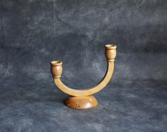 May include: A light-colored wooden candle holder with two candle cups. The candle holder has a curved design and sits on a round base. The wood grain is visible. The background is a mottled gray.