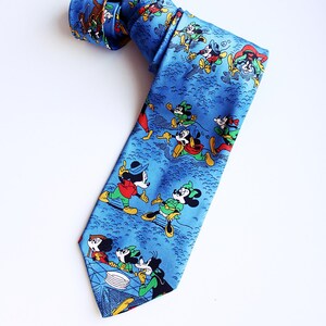 Könnte beinhalten: Blaue Krawatte mit einem sich wiederholenden Muster von Mickey Mouse und seinen Freunden in Winterkleidung. Die Krawatte ist aus glattem Stoff gefertigt und hat ein klassisches spitzes Ende. Das Design umfasst Charaktere in Rot, Grün und Schwarz.