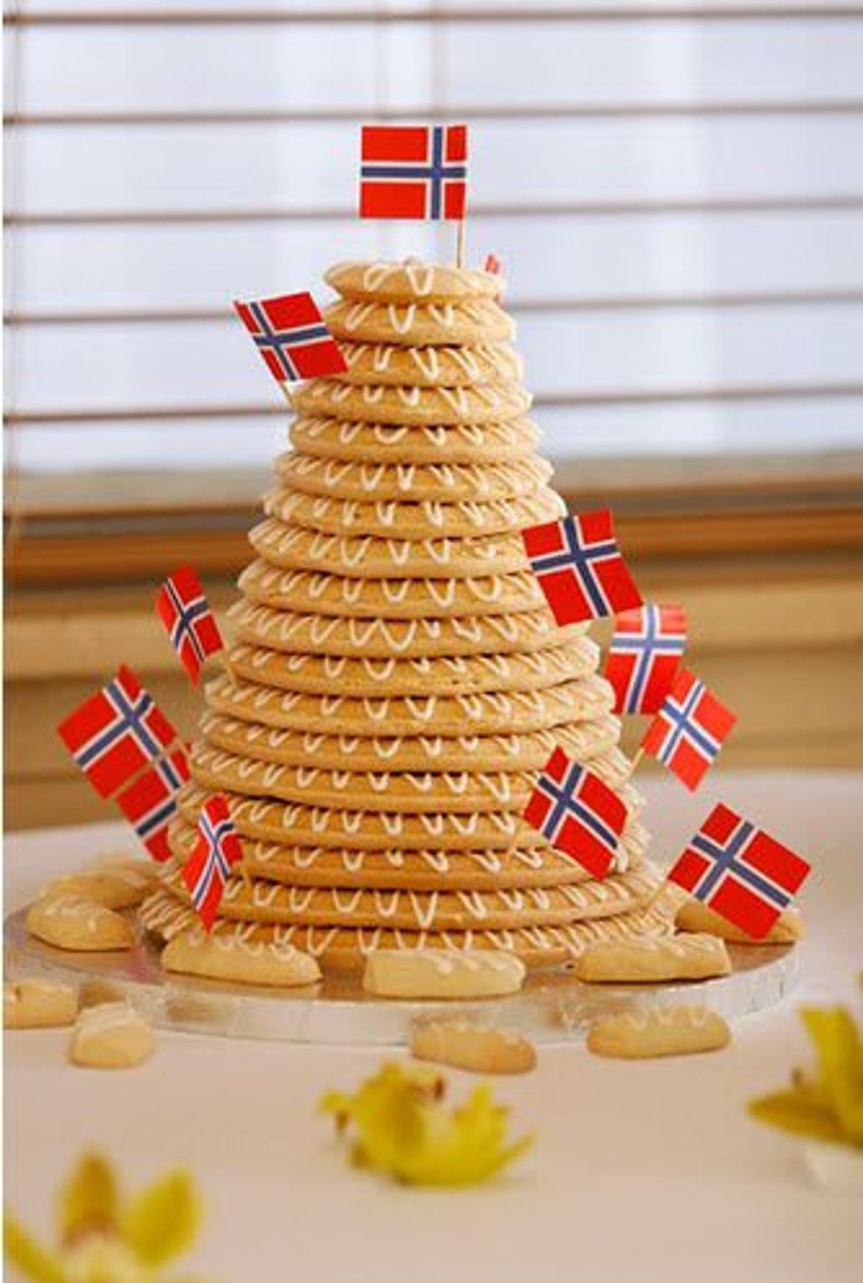 Puede incluir: Una torre de galletas de color marr&oacute;n dorado, decoradas con glaseado blanco, est&aacute; adornada con peque&ntilde;as banderas noruegas. Las galletas est&aacute;n dispuestas en forma c&oacute;nica, parecida a un postre festivo. Varias galletas est&aacute;n esparcidas en la base.