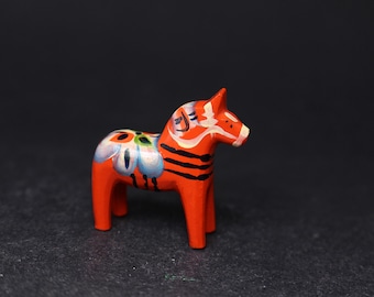May include: A small, hand-painted wooden Dala horse figurine. The horse is primarily orange with black stripes and floral designs in blue, white, and green. The traditional Swedish folk art piece is a decorative item.