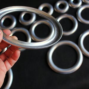 Puede incluir: Una colecci&oacute;n de anillos met&aacute;licos plateados de varios tama&ntilde;os. Un anillo ovalado se sostiene con la mano, con varios otros anillos circulares en el fondo. Los anillos tienen un acabado met&aacute;lico liso y est&aacute;n sobre un fondo oscuro.