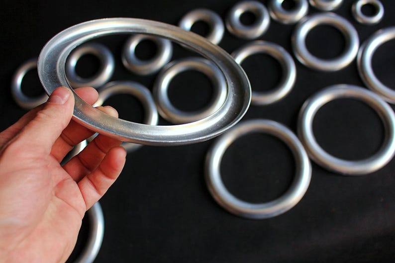 Puede incluir: Una colecci&oacute;n de anillos met&aacute;licos plateados de varios tama&ntilde;os. Un anillo ovalado se sostiene con la mano, con varios otros anillos circulares en el fondo. Los anillos tienen un acabado met&aacute;lico liso y est&aacute;n sobre un fondo oscuro.