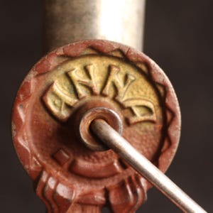 May include: Close-up of a vintage key with a circular, reddish-brown head. The head features the word "ANNA" in a gold-colored, stylized font. A silver-colored rod extends from the key head, suggesting a functional design. The key is likely for a lock.