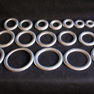 Puede incluir: Colecci&oacute;n de anillos met&aacute;licos plateados de varios tama&ntilde;os dispuestos sobre una superficie negra. Los anillos son circulares, con un grosor uniforme, y var&iacute;an desde formas ovaladas grandes hasta tama&ntilde;os peque&ntilde;os y redondos. Se destaca el brillo met&aacute;lico.