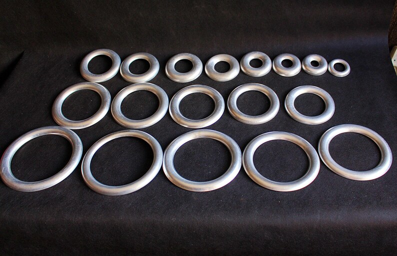 Puede incluir: Colecci&oacute;n de anillos met&aacute;licos plateados de varios tama&ntilde;os dispuestos sobre una superficie negra. Los anillos son circulares, con un grosor uniforme, y var&iacute;an desde formas ovaladas grandes hasta tama&ntilde;os peque&ntilde;os y redondos. Se destaca el brillo met&aacute;lico.