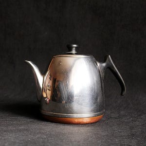 May include: A stainless steel teapot with a black handle and lid. The teapot has a wooden base and a curved spout. The teapot is a vintage style and is set against a dark background.