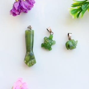 Op de afbeelding: Drie groene jade hangers met zilverkleurige beugels. Eén hanger is een vuist, een andere is een schildpad en de derde is een olifant. De hangers worden gepresenteerd op een wit oppervlak, met paarse en groene bloemaccenten.