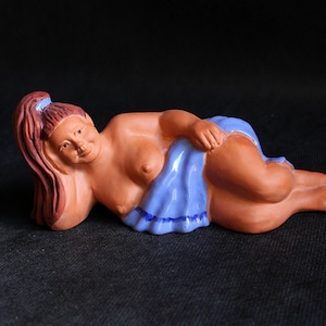 May include: A ceramic figurine of a reclining figure with brown skin, red hair, and a blue cloth. The figure is lying on its side, with one arm supporting its head. The blue cloth covers the lower body. The figurine is on a black surface.