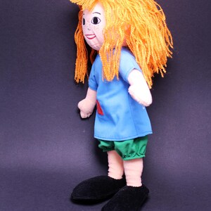 Pode incluir: Boneca feita &agrave; m&atilde;o com cabelo de fio laranja, uma camisa azul com um remendo vermelho, cal&ccedil;&otilde;es verdes e sapatos pretos. A boneca tem um rosto pintado com sardas, em um fundo escuro.