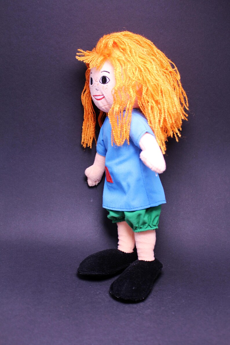 Pode incluir: Boneca feita &agrave; m&atilde;o com cabelo de fio laranja, uma camisa azul com um remendo vermelho, cal&ccedil;&otilde;es verdes e sapatos pretos. A boneca tem um rosto pintado com sardas, em um fundo escuro.