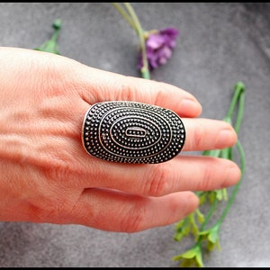 Peut inclure: Une bague ovale argentée avec une surface texturée ornée de petits points en relief. La bague est portée sur un doigt, sur un fond gris avec des brins de verdure et des fleurs violettes.