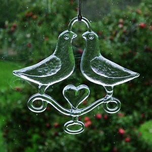 Puede incluir: Un adorno de vidrio con dos palomas con un coraz&oacute;n en el centro, colgado de una cuerda. Las palomas se miran y tienen un acabado de vidrio transparente.