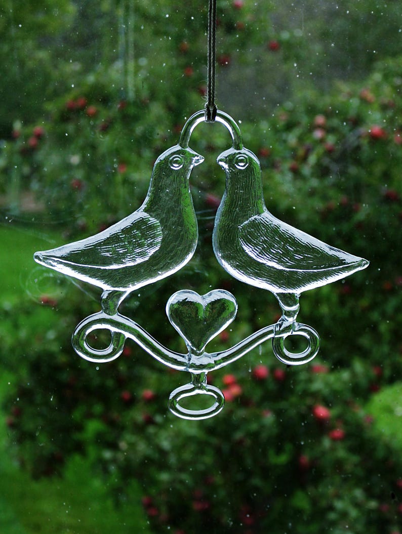 Puede incluir: Un adorno de vidrio con dos palomas con un coraz&oacute;n en el centro, colgado de una cuerda. Las palomas se miran y tienen un acabado de vidrio transparente.