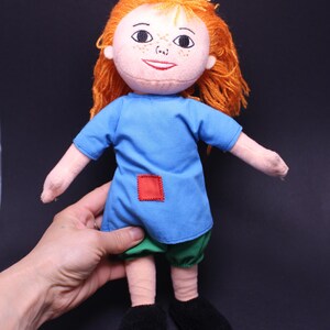 Pode incluir: Boneca feita &agrave; m&atilde;o com cabelo de fio laranja, vestido azul, cal&ccedil;&otilde;es verdes e sapatos pretos. A boneca tem um rosto costurado com sardas e um remendo quadrado vermelho no vestido. A boneca tem aproximadamente 25 cm de altura.