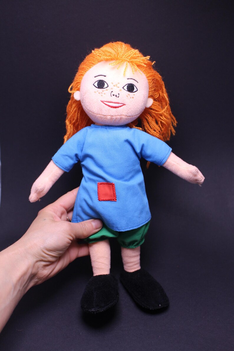 Pode incluir: Boneca feita &agrave; m&atilde;o com cabelo de fio laranja, vestido azul, cal&ccedil;&otilde;es verdes e sapatos pretos. A boneca tem um rosto costurado com sardas e um remendo quadrado vermelho no vestido. A boneca tem aproximadamente 25 cm de altura.