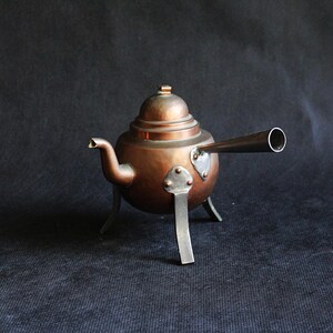 Op de afbeelding: Een koperkleurige theepot met een tuit en een lange, cilindrische pijp die zich vanaf de zijkant uitstrekt. De theepot staat op drie kleine pootjes en heeft een deksel. Het metaal heeft een warme, roodbruine tint.