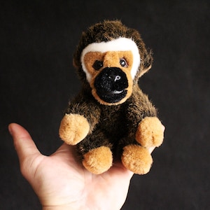 Puede incluir: Un mono de peluche con pelaje marrón, manos y pies color canela, y una franja blanca en la frente. El mono tiene una cara negra y se sostiene en una mano sobre un fondo negro. El juguete es un peluche.
