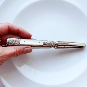 Op de afbeelding: Een zilveren en parelmoeren zakmes met sierlijke details. Het mes is open en toont een glanzend lemmet. De handgreep heeft een wit, iriserend materiaal. Het mes ligt op een witte plaat.