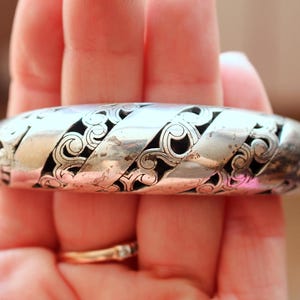 Puede incluir: Un brazalete plateado con un intrincado dise&ntilde;o calado. El brazalete presenta un patr&oacute;n repetitivo de motivos arremolinados en forma de volutas, con espacios oscuros entre el trabajo en metal. Es una pieza vintage.