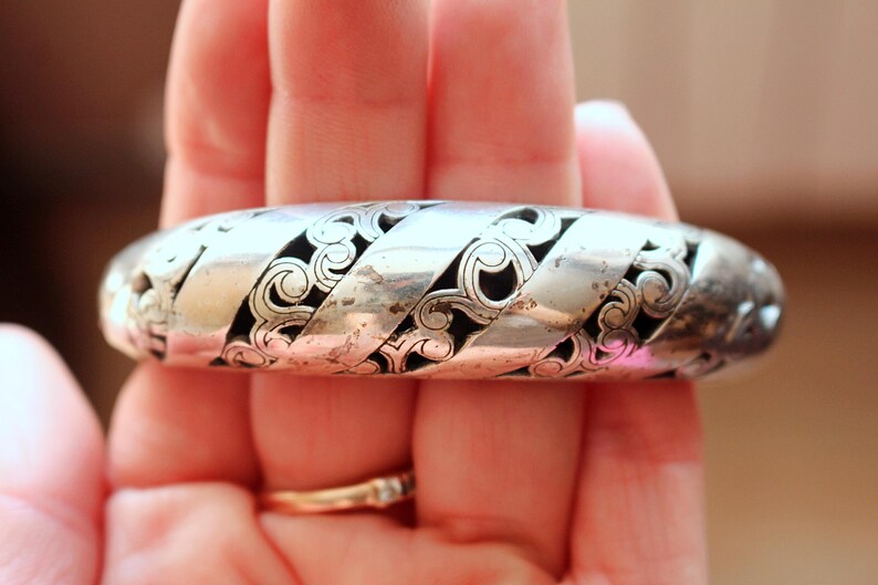Puede incluir: Un brazalete plateado con un intrincado dise&ntilde;o calado. El brazalete presenta un patr&oacute;n repetitivo de motivos arremolinados en forma de volutas, con espacios oscuros entre el trabajo en metal. Es una pieza vintage.