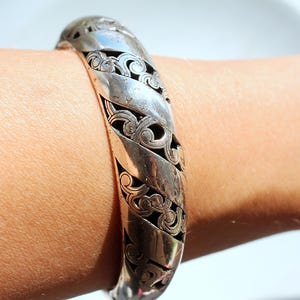 Puede incluir: Un brazalete plateado con un intrincado dise&ntilde;o de filigrana arremolinada. El brazalete se lleva en la mu&ntilde;eca y presenta una serie de bandas curvas y texturizadas. El dise&ntilde;o calado revela un interior oscuro, creando un contraste con el metal brillante.