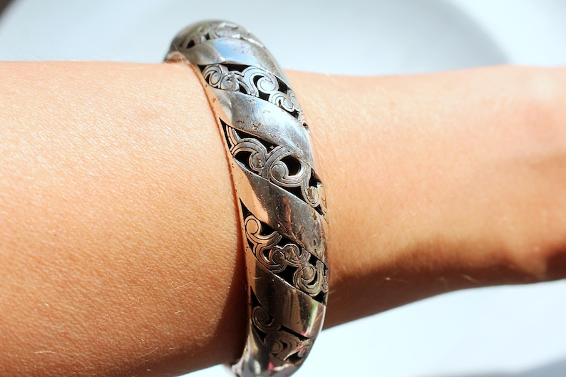 Puede incluir: Un brazalete plateado con un intrincado dise&ntilde;o de filigrana arremolinada. El brazalete se lleva en la mu&ntilde;eca y presenta una serie de bandas curvas y texturizadas. El dise&ntilde;o calado revela un interior oscuro, creando un contraste con el metal brillante.