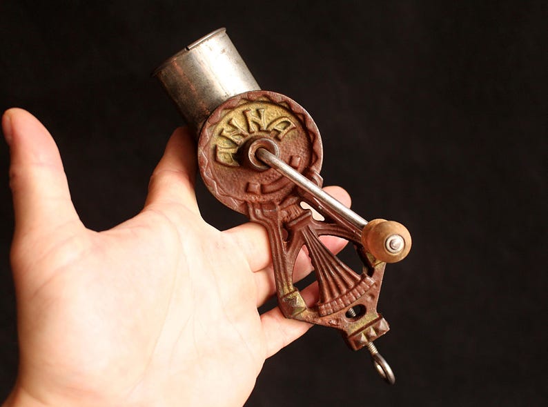 May include: Antique hand-crank coffee grinder with a brown and gold finish. The grinder features a silver hopper and a wooden handle. The word "KING" is embossed on the grinder's wheel. A vintage kitchen tool.