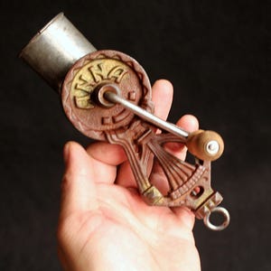 May include: Antique hand-cranked food grinder with a brown and gold cast iron body and a silver metal hopper. The grinder features a wooden handle and a circular design with the word "INA" on it.