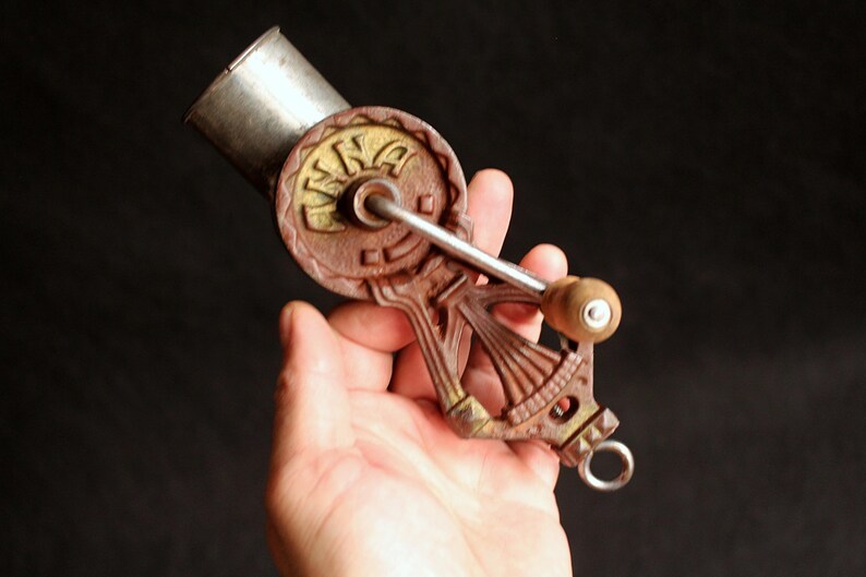 May include: Antique hand-cranked food grinder with a brown and gold cast iron body and a silver metal hopper. The grinder features a wooden handle and a circular design with the word "INA" on it.