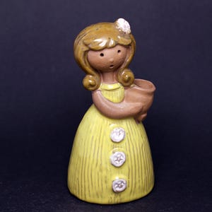 Może przedstawiać: Ceramiczna figurka dziewczynki w żółtej sukience z pionowymi liniami i trzema białymi guzikami. Dziewczynka ma brązowe włosy, mały biały kwiat i trzyma brązowy garnek. Figurka na ciemnym tle.