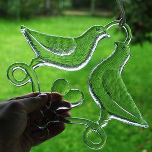 Puede incluir: Un adorno de vidrio con dos palomas posadas en una rama con un dise&ntilde;o en forma de coraz&oacute;n. Las palomas son de vidrio transparente y la rama es de color marr&oacute;n claro.