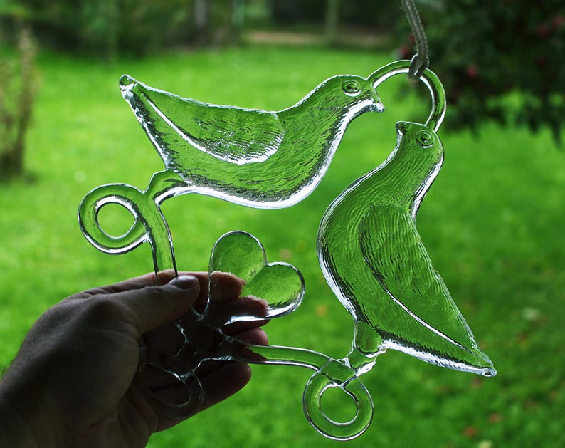 Puede incluir: Un adorno de vidrio con dos palomas posadas en una rama con un dise&ntilde;o en forma de coraz&oacute;n. Las palomas son de vidrio transparente y la rama es de color marr&oacute;n claro.