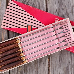 Può includere: Un set da fonduta vintage con sei forchette, ciascuna con manico in legno e rebbi in acciaio inossidabile. Il set è presentato in una scatola rosa e rossa con un design a strisce, ideale per le feste.