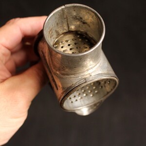 May include: Vintage metal kitchen tool with a cylindrical top and a perforated base. The tool is made of a silver-colored metal and has a T-shape. The tool is held in a hand against a dark background.
